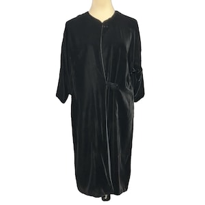 May include: A long-sleeved black velvet duster coat featuring a smooth, soft-touch fabric texture, a simple round neckline, and a single circular button closure positioned at the waistline, creating a clean, straight-cut silhouette.