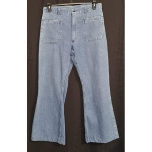 May include: Light blue denim pants with two large front pockets and a button closure. The pants have a flared leg design. The pants are hanging on a black hanger against a dark background.