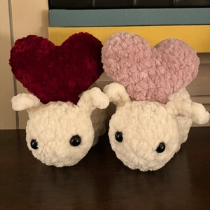 May include: Two white plush toys with large black eyes. Each toy has a heart-shaped plush on top, one is red and the other is pink.