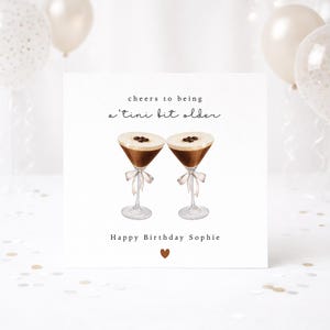 May include: A birthday card featuring two watercolour martini glasses filled with a dark brown liquid and coffee beans. The card reads "cheers to being a 'tini bit older" and "Happy Birthday Sophie". A small brown heart is at the bottom.