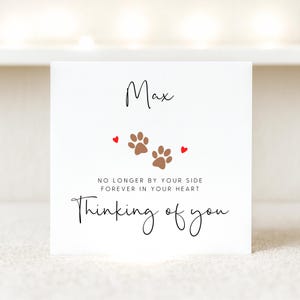 A large white square  lightly textured card, where a name can be added, along with the words 'No longer by your side, forever in your heart, thinking of you'. The card features a paw print and love heart simple illustration.