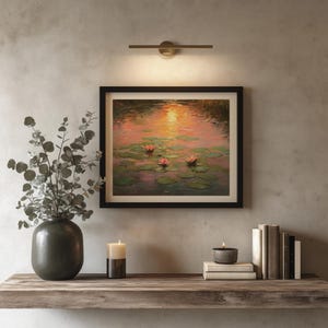 May include: A framed painting of water lilies in a pond, with a black frame, hangs above a wooden shelf. A dark green vase with leafy branches, a candle, and books are on the shelf. A brass light fixture is above the painting.