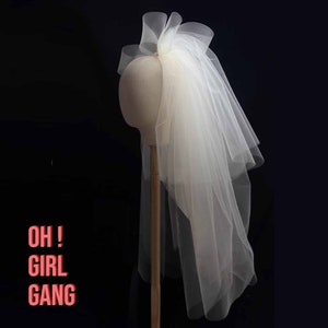 May include: A white tulle bridal veil with a large bow at the top. The veil is draped over a mannequin head and is displayed against a black background.