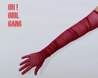 Può includere: Un paio di lunghi guanti in rete rossa. I guanti sono raccolti al polso e si estendono fino al gomito. Il testo "OH! GIRL GANG" è scritto in lettere rosa su uno sfondo bianco.