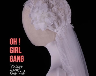 May include: A vintage lace cap veil with a large white flower and delicate lace detailing. The veil is made of sheer white fabric and is displayed on a mannequin head. Text on the image reads "OH! GIRL GANG" and "Vintage Lace Cap Veil."
