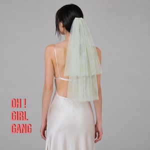 Op de afbeelding: Een lichtgroene, gelaagde tule sluier is bevestigd aan de achterkant van het hoofd van een persoon. De persoon draagt een witte satijnen jurk met een lage rug. De woorden "OH! GIRL GANG" zijn roze gedrukt aan de linkerkant van de afbeelding.
