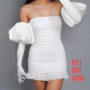 Op de afbeelding: Een witte off-shoulder jurk met grote, pofmouwen en een aansluitend lijfje. De jurk heeft een gerimpelde zoom en de tekst "OH! GIRL GANG" is afgedrukt in de rechterbenedenhoek van de afbeelding.