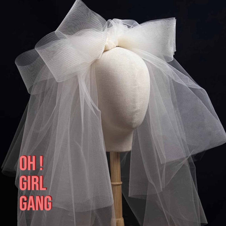May include: A white tulle bridal veil with a large bow on top. The veil is on a white mannequin head. The text "OH! GIRL GANG" is written in pink letters on the bottom of the image.