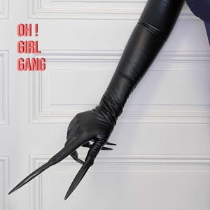 May include: A black leather glove with long, sharp, black claws. The glove is extended out in front of a white wall with the text "OH! GIRL GANG" written in pink.