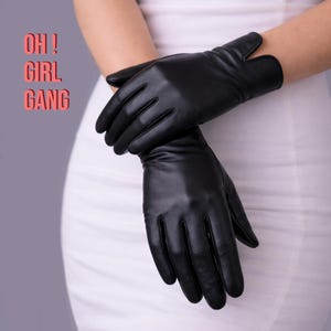 Może przedstawiać: Para czarnych skórzanych rękawiczek. Rękawiczki są gładkie i dopasowane, o klasycznym kroju. Tekst "OH! GIRL GANG" jest w różowych literach w lewym górnym rogu.