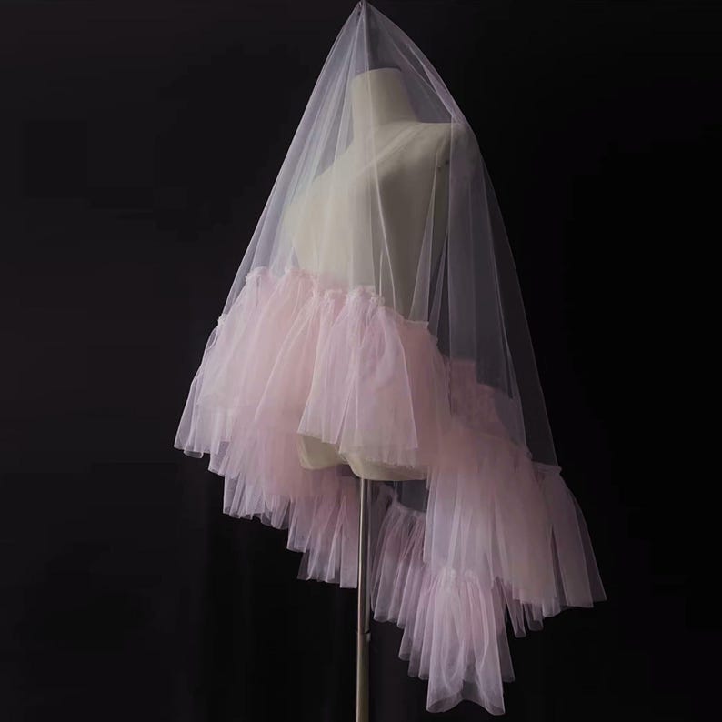 Puede incluir: Un velo de novia con una falda con volantes en un tono rosa suave. El velo est&aacute; hecho de tela blanca transparente que cae sobre un maniqu&iacute;. La falda presenta m&uacute;ltiples capas de tul delicado y con volantes, creando un aspecto rom&aacute;ntico.