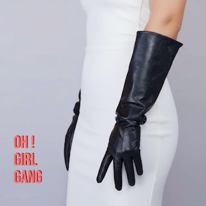 Könnte beinhalten: Schwarze Leder-Opernhandschuhe. Die Handschuhe reichen über den Ellbogen hinaus. Der Text "OH! GIRL GANG" befindet sich in der unteren linken Ecke des Bildes. Die Handschuhe werden mit einem weißen Kleid getragen.