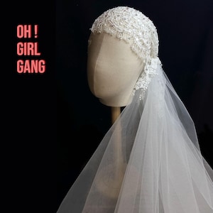 Peut inclure: Un voile de mariée blanc avec un coiffe orné de perles et de dentelle. Le voile est fait d'un tissu transparent et fluide. Le texte "OH ! GIRL GANG" est en lettres roses sur le côté gauche de l'image.