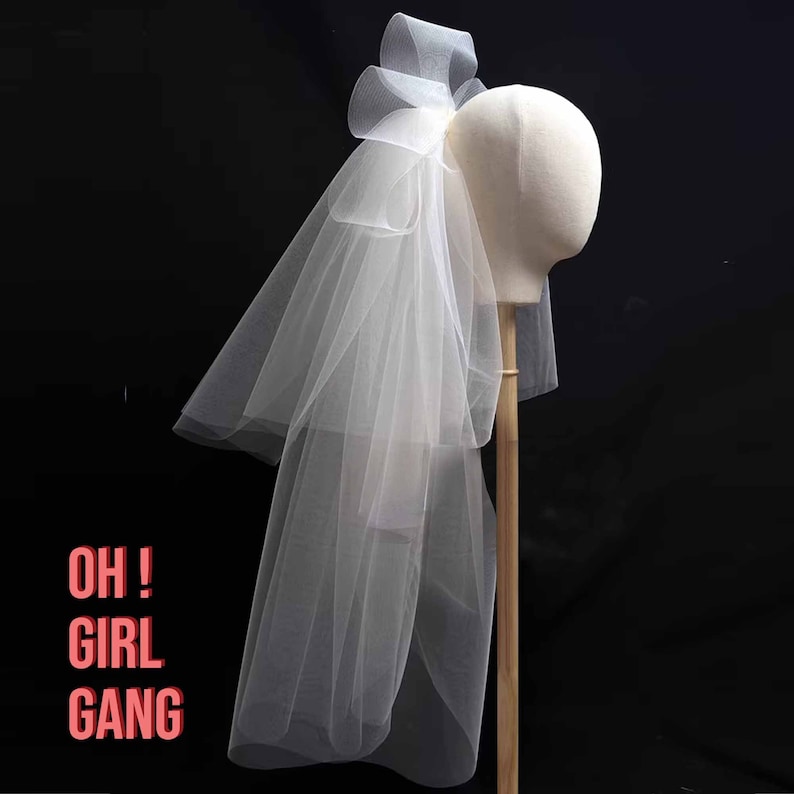 May include: A white tulle bridal veil with a large bow at the top. The veil is displayed on a mannequin head.