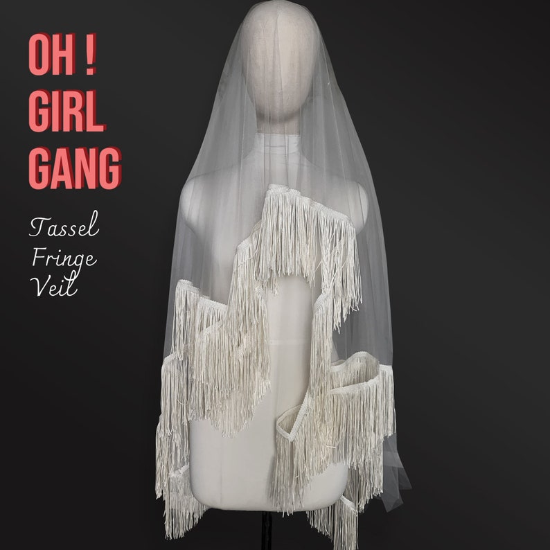 Może przedstawiać: Biały welon tiulowy z frędzlami. Welon znajduje się na białym manekinie. Tekst "OH! GIRL GANG" jest czerwonymi literami na czarnym tle. Tekst "Tassel Fringe Veil" jest czarnymi literami na białym tle.