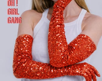 Peut inclure: Une paire de longs gants d'opéra à sequins rouge vif qui dépassent les coudes. Les gants sont recouverts de petites paillettes rouges réfléchissantes. Le texte "OH! GIRL GANG" est imprimé en police orange-rouge sur le côté gauche.