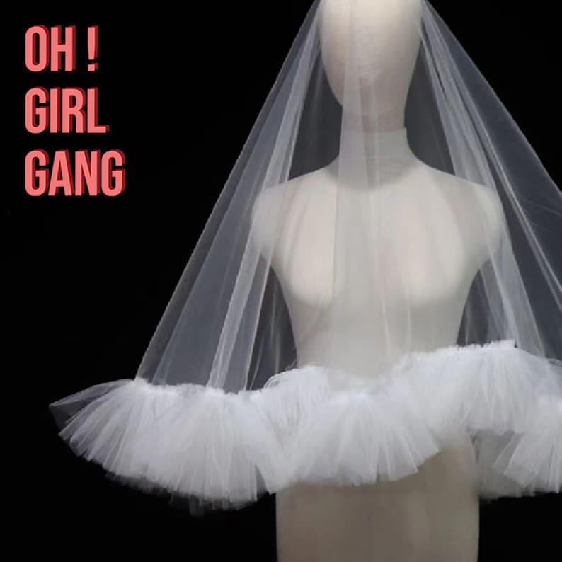 Puede incluir: Un velo de novia de tul blanco con un ribete con volantes, exhibido en un maniqu&iacute;. El texto "OH! GIRL GANG" est&aacute; en letras rosas en la esquina superior izquierda. El velo es transparente y delicado, ideal para una boda.