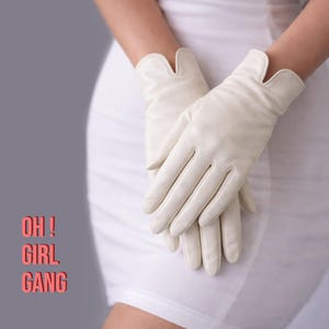 Könnte beinhalten: Cremefarbene Lederhandschuhe mit einem einzigartigen, gewellten Randdesign. Die Handschuhe werden vor einem weißen Kleid und einem grauen Hintergrund gezeigt. Der Text "OH! GIRL GANG" ist rot dargestellt.