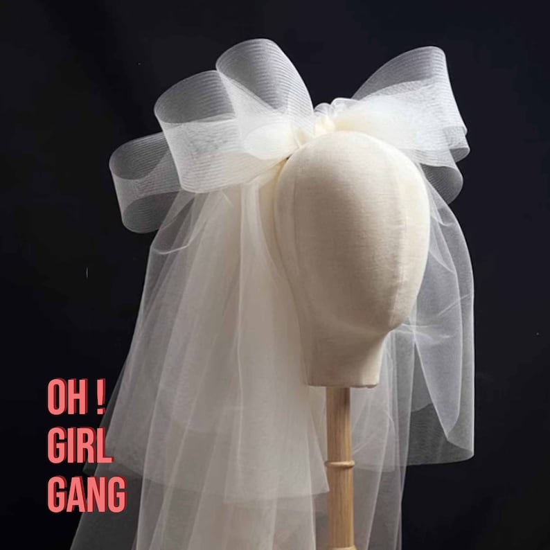 May include: A white tulle veil with a large bow on top. The veil is on a mannequin head. The text "OH! GIRL GANG" is written in pink on the veil.