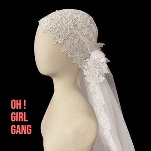 Peut inclure: Un voile de mariée en dentelle blanche avec des motifs floraux et de feuilles est présenté sur une tête de mannequin. Le voile est fait de dentelle délicate et de tissu transparent. Le texte "OH ! GIRL GANG" est dans le coin inférieur gauche.