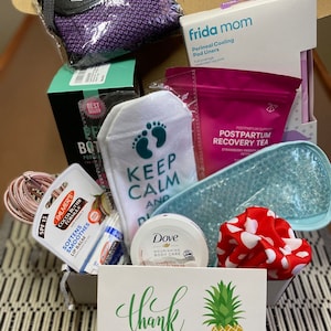 May include: A gift box filled with various items, including a cooling towel, a pair of socks with the text "Keep Calm and Drink", a bottle of postpartum recovery tea, a tube of lip balm, a jar of body cream, a cooling pad, and a thank you card with a pineapple design.