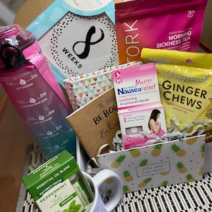 May include: A gift box filled with pregnancy-related items, including a white mug with the text "Proud Mom Advocate", a small bottle of peppermint essential oil, a pack of ginger chews, a pack of acupressure bands for nausea relief, a notebook with the text "Be Bold Be Brave Be Human", and a pink water bottle with a motivational quote.