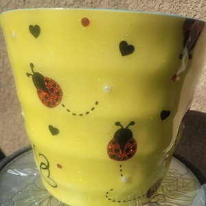 Puede incluir: Una maceta de cerámica amarilla brillante decorada con mariquitas, corazones y líneas de puntos. Las mariquitas tienen detalles rojos y negros. Pequeñas perlas blancas y puntos rojos también están dispersos.