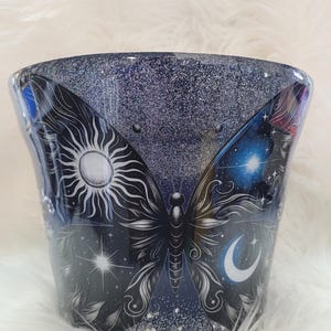 May include: A cylindrical tumbler featuring a dark blue glitter finish and a large butterfly graphic. The butterfly wings display celestial imagery, including a white sun on the left wing and a crescent moon with stars on the right wing.