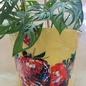 May include: A yellow, glitter-covered plant pot featuring a mosaic strawberry design. The pot contains a green plant with distinctive, hole-filled leaves. The plant is suspended by a hanging wire.