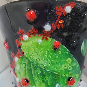 May include: A black cylindrical planter decorated with a vibrant green leaf motif covered in realistic water droplets. The design features several small red ladybugs with black spots and delicate red vine accents with tiny white floral embellishments.