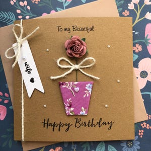 May include: A square brown greeting card featuring a paper rose, a floral patterned pot, and a twine bow. The text reads To my Beautiful and Happy Birthday. A white tag attached with twine displays the word wife and a small black heart icon.