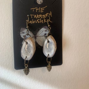 May include: A pair of earrings featuring a white antler design and a small pine cone charm. The earrings are displayed on a black card with the text "THE Tattooed Babushka".