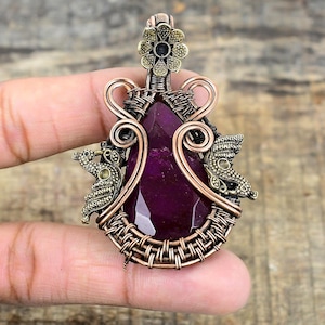 Puede incluir: Un colgante de piedra preciosa púrpura con envoltura de alambre de cobre y detalles dorados. El colgante presenta un diseño de flor en la parte superior y dos figuras de pájaros en los lados.