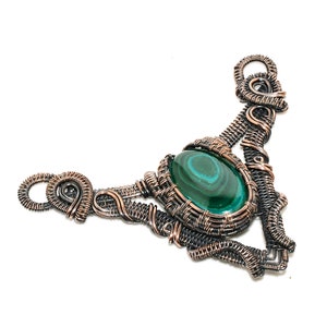 Puede incluir: Un collar artesanal de alambre de cobre con una piedra ovalada de malaquita. La piedra verde está engastada en un marco de alambre tejido, con intrincados trabajos de alambre que se extienden a los lados. El collar tiene un diseño artístico único.