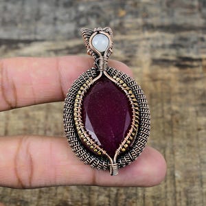 Puede incluir: Colgante artesanal con una piedra preciosa roja oscura de forma ovalada. La piedra está engastada en un intrincado marco de alambre de cobre, con detalles de metal dorado y una pequeña piedra blanca en la parte superior. Diseñado para ser usado como collar.