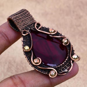 May include: A dark red gemstone pendant with a copper wire frame and gold accents. The pendant is shaped like a teardrop and has a detailed wire design.