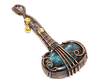 Puede incluir: Colgante artesanal con forma de instrumento de cuerda. El colgante presenta una gran piedra de labradorita ovalada con iridiscencia azul y verde, envuelta en alambre de cobre. Cuentas doradas están unidas al mango.