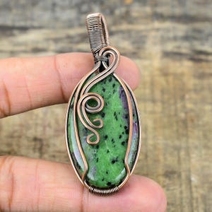 May include: A green and black gemstone pendant with copper wire wrapping. The pendant is oval shaped and has a swirl design in the wire.