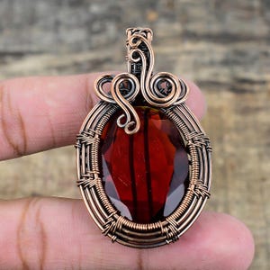 May include: A copper wire wrapped pendant featuring a red gemstone. The pendant has a detailed, ornate design with a bail at the top for attaching a chain.