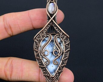 Peut inclure: Pendentif artisanal en fil de cuivre avec une pierre en forme de goutte d'eau, bleu clair et blanche. Le pendentif présente un motif en fil tissé et une petite pierre ovale en haut. Le pendentif est tenu à la main sur fond noir.