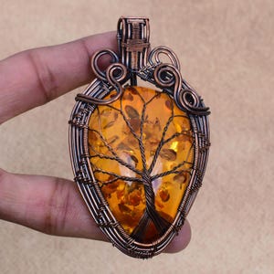 May include: A copper wire wrapped pendant featuring a tree of life design with a large amber stone. The stone is a deep orange colour with inclusions of smaller amber pieces.