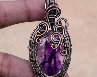 May include: A purple amethyst gemstone pendant wrapped in copper wire with intricate designs. The pendant is oval shaped and has a copper bail for hanging.
