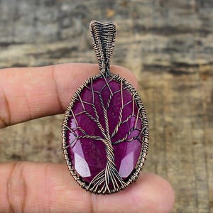 May include: Oval-shaped pendant with a deep red gemstone, featuring a wire-wrapped tree design. The pendant has a copper-colored metal frame and bail. The gemstone is polished and reflects light, creating a striking contrast with the darker metal.