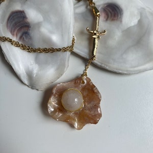 Peut inclure: Collier doré avec un pendentif orné d'une pierre précieuse rose clair dans un cadre doré. Le pendentif est attaché à une chaîne dorée avec un charm en forme d'épée décorative.
