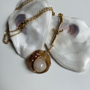 Può includere: Una collana color oro con un ciondolo con una pietra rosa chiaro incastonata in una cornice marrone e oro. La collana ha anche un piccolo ciondolo a forma di spada. La collana è esposta su una superficie bianca con due conchiglie.
