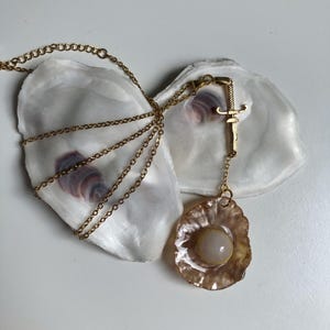 Peut inclure: Un collier doré avec un pendentif orné d'une pierre de couleur claire sertie dans un cadre texturé doré. Le collier est présenté sur une surface blanche avec deux coquilles d'huîtres ouvertes.