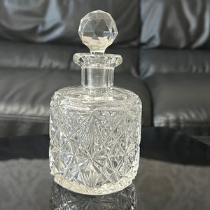 Puede incluir: Una botella de vidrio transparente con un tapón facetado. La botella tiene un diseño de vidrio cortado y está sobre una superficie negra.