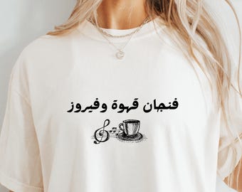 May include: Ivory-colored t-shirt with black Arabic text and a musical note design featuring a coffee cup. The text reads "Fanjan Qahwa Wa Fairouz". The shirt is a casual, comfortable style.