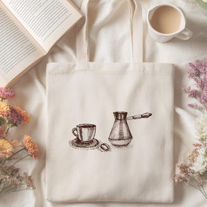 Può includere: Borsa tote color crema con un'illustrazione a inchiostro di una tazza di caffè, un piattino, un chicco di caffè e una caffettiera tradizionale. La borsa è circondata da un libro, fiori e una tazza di caffè, creando una scena accogliente.