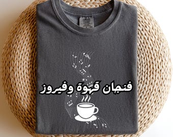 May include: Pepper-colored t-shirt with a white graphic of a coffee cup and musical notes. The Arabic text on the shirt reads "Coffee and Vibes". The shirt is displayed on a woven, circular surface.
